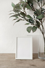 Empty picture frame mockup standing on wooden tabletop with green floral bouquet, white wall...