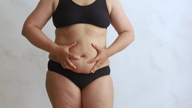 Unrecognizable obese woman wearing black underwear, squeezing excessive subcutaneous fat of abdomen, doing massage on white background. Weight loss, adiposity, plastic surgery, liposuction, obesity. 