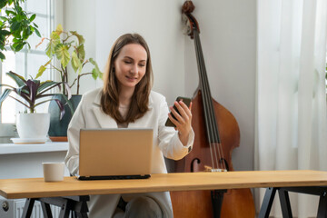 Woman is sitting at her desk, holding a mobile phone, staring at a laptop, synchronizing data...