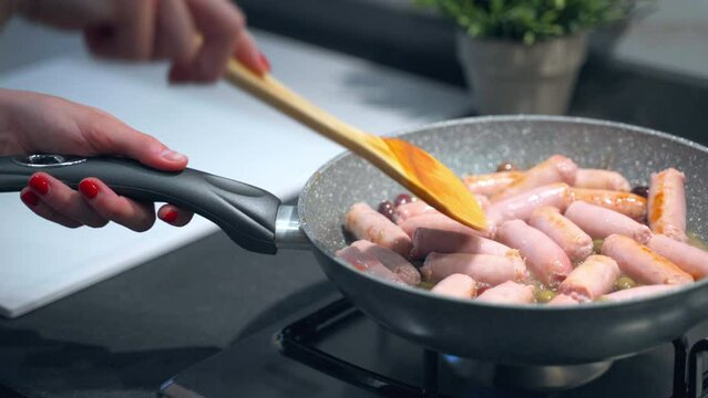 Woman cooking on trendy gas stove in the kitchen, chef frying cut sausages in pan with extra virgin olive oil and olives mixing with wooden spoon. Preparation of fat greasy sausages on frying pan