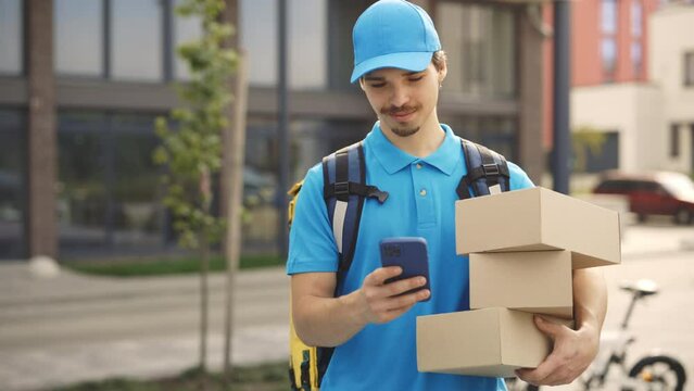 Smiling Courier Going Along Building Holding the Paper Boxes and Mobile Phone in Hand. Delivery Man Looking for Delivery Address on Street. Handsome Guy Using Smartphone While Delivering Outdoors