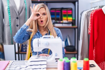 Blonde woman dressmaker designer using sew machine doing ok gesture shocked with surprised face, eye looking through fingers. unbelieving expression.