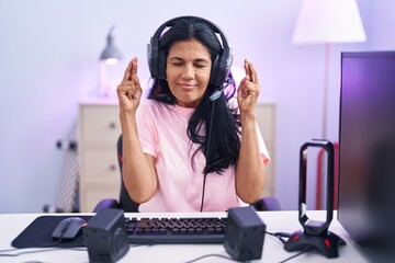 Mature hispanic woman playing video games at home gesturing finger crossed smiling with hope and eyes closed. luck and superstitious concept.