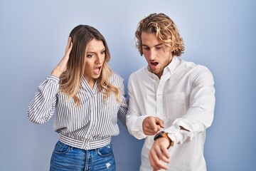 Young couple standing over blue background in hurry pointing to watch time, impatience, upset and...