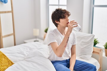 Young hispanic man drinking glass of water sitting on bed at bedroom