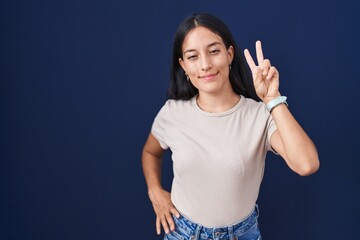 Young hispanic woman standing over blue background smiling looking to the camera showing fingers doing victory sign. number two.