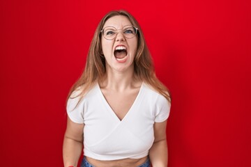 Young caucasian woman standing over red background angry and mad screaming frustrated and furious,...