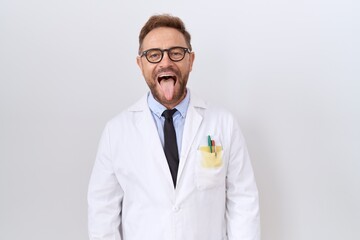 Middle age doctor man with beard wearing white coat sticking tongue out happy with funny expression. emotion concept.