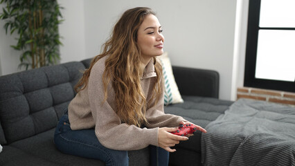 Young beautiful hispanic woman playing video game sitting on sofa at home