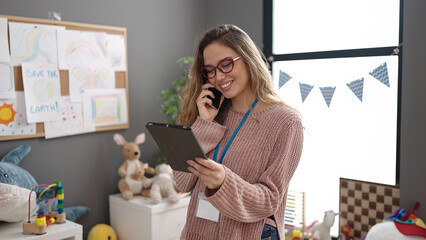 Young beautiful hispanic woman preschool teacher talking on smartphone using touchpad at kindergarten