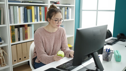 Young beautiful hispanic woman student using computer eating apple at library university
