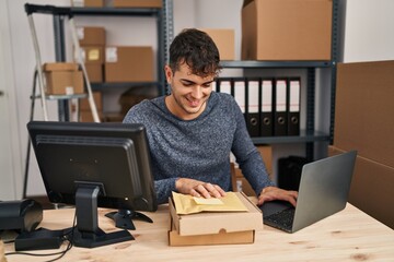 Young hispanic man ecommerce business worker using laptop holding packages at office