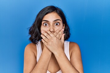 Young hispanic woman standing over blue background shocked covering mouth with hands for mistake....