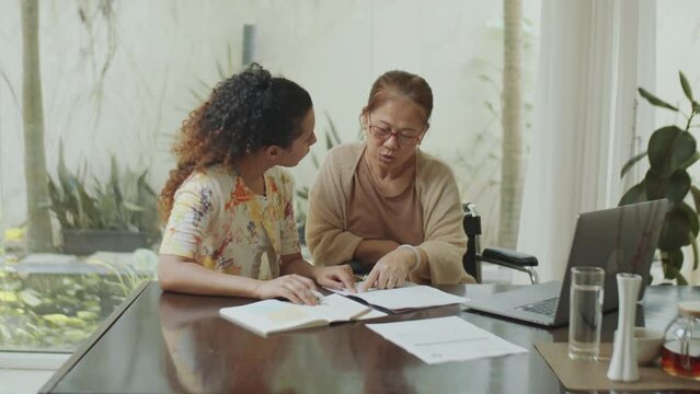 Young female financial advisor using laptop and explaining documents to senior Asian woman in wheelchair during wealth management consultation at home