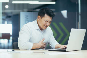 An office worker of Asian origin is working on a laptop and holding his chest with his hand. Suffering and writhing in pain, he needs medical help.