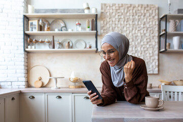 Pleasantly surprised young Muslim woman in hijab at home in the kitchen looking at the phone with admiration. Received money, read good news, messages. Shows a victory gesture with his hand.