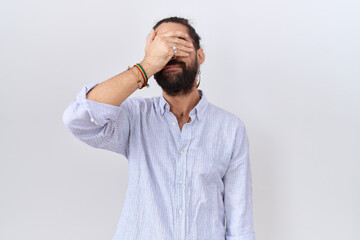 Hispanic man with beard wearing casual shirt covering eyes with hand, looking serious and sad....