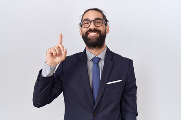 Hispanic man with beard wearing suit and tie showing and pointing up with finger number one while smiling confident and happy.