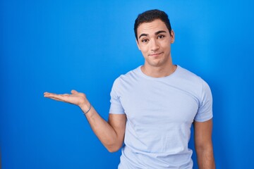 Fototapeta premium Young hispanic man standing over blue background smiling cheerful presenting and pointing with palm of hand looking at the camera.