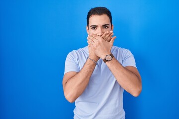 Young hispanic man standing over blue background shocked covering mouth with hands for mistake. secret concept.