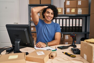 Hispanic man with curly hair working at small business ecommerce smiling confident touching hair with hand up gesture, posing attractive and fashionable