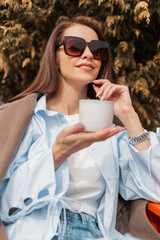 Beautiful fashionable young girl in trendy stylish clothes with sunglasses sits in a cafe outdoors and drinks tea