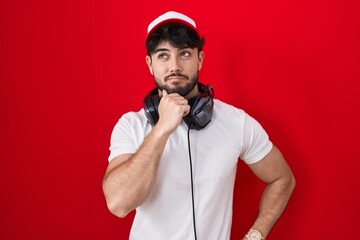 Hispanic man with beard wearing gamer hat and headphones with hand on chin thinking about question, pensive expression. smiling and thoughtful face. doubt concept. © Krakenimages.com