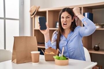 Young brunette woman eating take away food at home showing smartphone screen annoyed and frustrated shouting with anger, yelling crazy with anger and hand raised