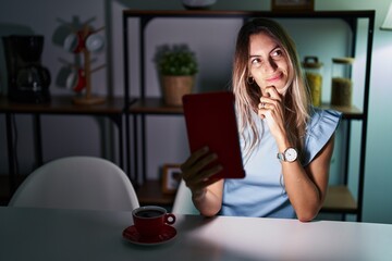 Young hispanic woman using touchpad sitting on the table at night with hand on chin thinking about question, pensive expression. smiling with thoughtful face. doubt concept.