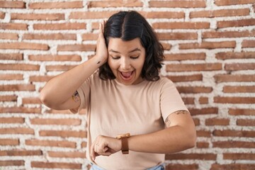 Young hispanic woman standing over bricks wall looking at the watch time worried, afraid of getting...