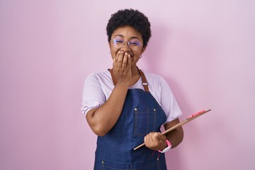 Young african american woman wearing professional waitress apron holding clipboard laughing and...