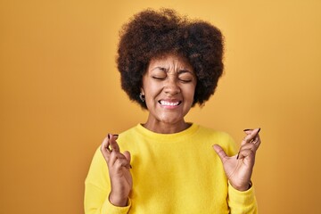 Young african american woman standing over yellow background gesturing finger crossed smiling with hope and eyes closed. luck and superstitious concept.