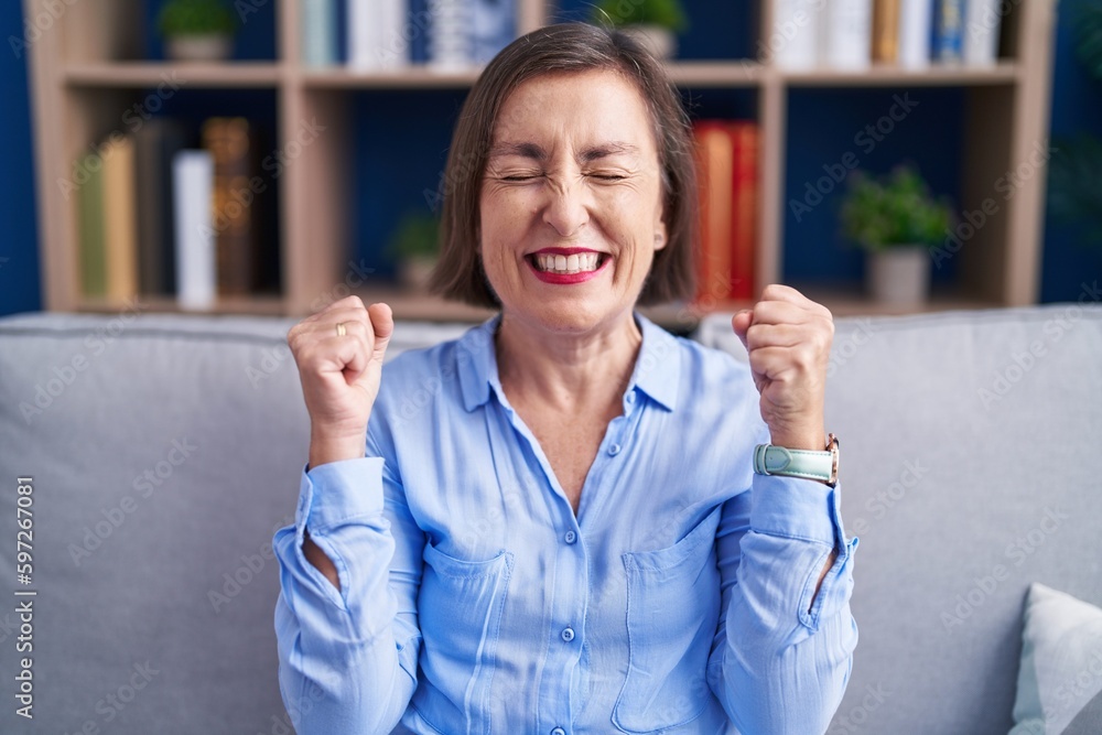 Wall mural Middle age hispanic woman sitting on the sofa at home excited for success with arms raised and eyes closed celebrating victory smiling. winner concept.