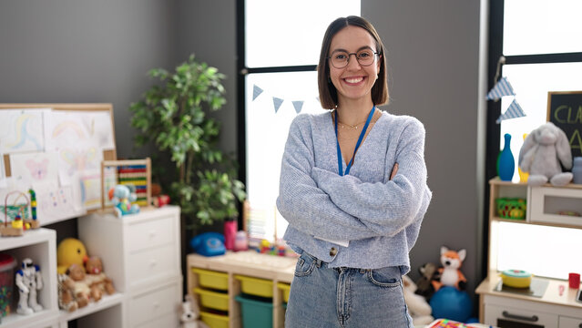 Young beautiful hispanic woman teacher smiling confident standing with arms crossed gesture at kindergarten