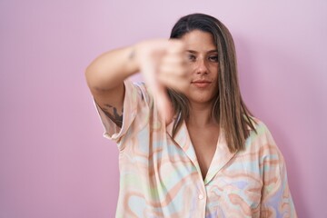 Blonde woman standing over pink background looking unhappy and angry showing rejection and negative with thumbs down gesture. bad expression.