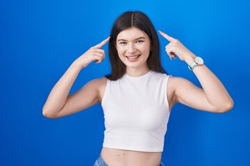 Fototapeta na wymiar Young caucasian woman standing over blue background smiling pointing to head with both hands finger, great idea or thought, good memory