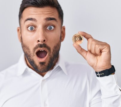 Handsome hispanic man holding cardano cryptocurrency coin scared and amazed with open mouth for surprise, disbelief face