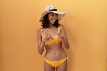 Young hispanic woman wearing bikini and summer hat pointing fingers to camera with happy and funny...