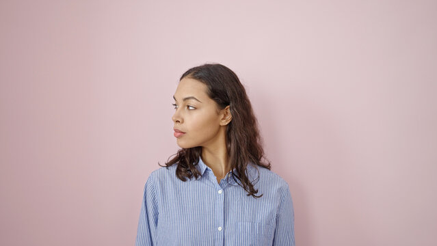 Young beautiful hispanic woman standing with serious expression looking to the side over isolated pink background