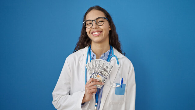 Young beautiful hispanic woman doctor smiling holding money over isolated blue background