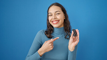 Young beautiful hispanic woman smiling confident pointing to key of new car over isolated blue...