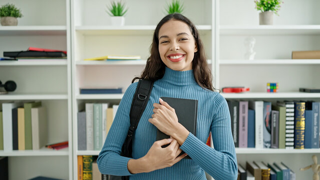 Young beautiful hispanic woman student standing holding book at university classroom