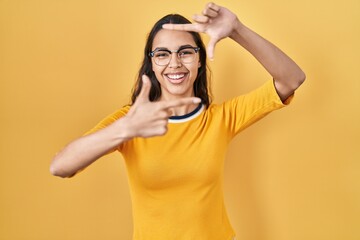 Fototapeta premium Young brazilian woman wearing glasses over yellow background smiling making frame with hands and fingers with happy face. creativity and photography concept.