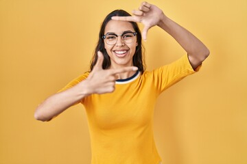 Young brazilian woman wearing glasses over yellow background smiling making frame with hands and fingers with happy face. creativity and photography concept.