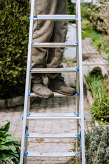 A worker, an adult male, a builder in a dirty robe stands on the steps of a metal, aluminum ladder in the open air. Photography, portrait of legs, shoe close-up, lifestyle, outdoor work.