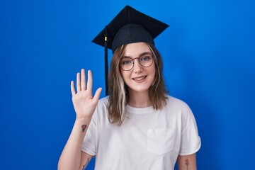 Blonde caucasian woman wearing graduation cap showing and pointing up with fingers number five while smiling confident and happy.