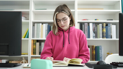 Young blonde woman student reading book sitting on table at library university