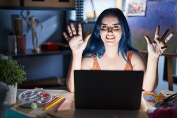 Young modern girl with blue hair sitting at art studio with laptop at night showing and pointing up...