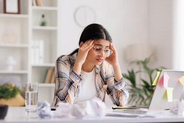 A tired young Indian girl gets angry from the number of tasks, crumples up the paper and throws it away, tries to calm down by meditating. Stressful work and study, overtasking, headache.
