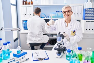 Middle age woman working at scientist laboratory smiling happy and positive, thumb up doing excellent and approval sign